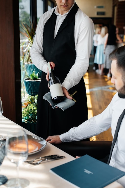 A young sommelier in a stylish uniform demonstrates and offers the client fine wine in the restaurant Customer service Table setting in a fine restaurant