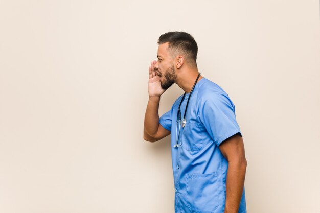 Young south-asian nurse man shouting and holding palm near opened mouth.