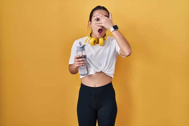 Young south asian woman wearing sportswear drinking water peeking in shock covering face and eyes with hand looking through fingers with embarrassed expression