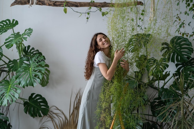 Young spanish girl stand among green plants on stepladder and hugging flower in living room hobbies