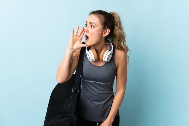 Young sport Brazilian woman with sport bag isolated on blue shouting with mouth wide open to the side