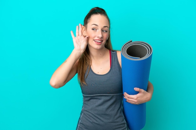 Young sport caucasian woman going to yoga classes while holding a mat isolated on blue background listening to something by putting hand on the ear