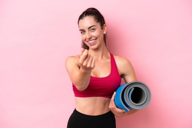 Young sport caucasian woman going to yoga classes while holding a mat isolated on pink background making money gesture