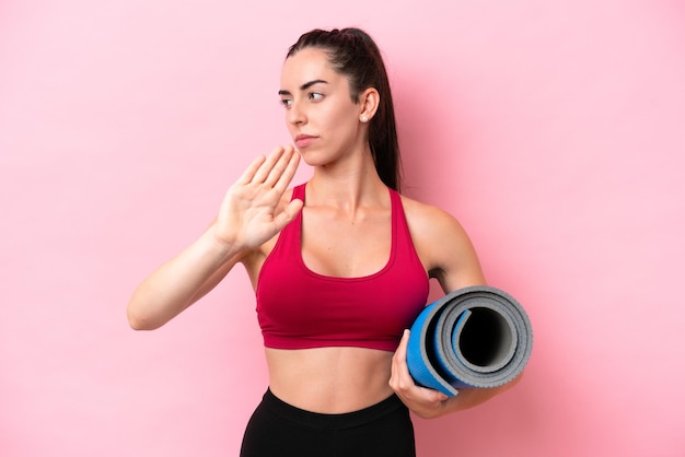 Young sport caucasian woman going to yoga classes while holding a mat isolated on pink background making stop gesture and disappointed