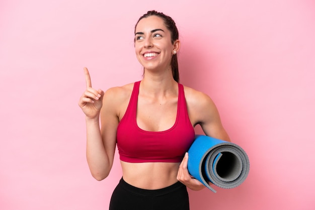 Young sport caucasian woman going to yoga classes while holding a mat isolated on pink background pointing up a great idea