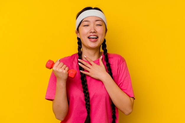 Young sport chinese woman isolated on yellow laughs out loudly keeping hand on chest.