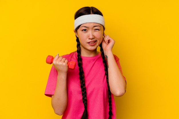 Young sport chinese woman isolated on yellow wall covering ears with hands