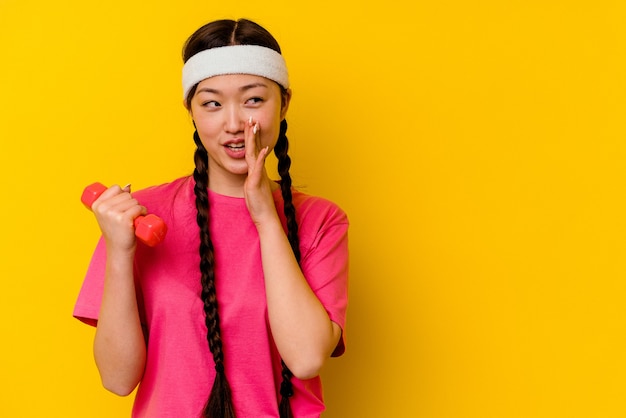 Young sport chinese woman isolated on yellow wall is saying a secret hot braking news and looking aside