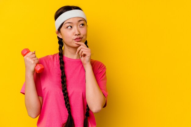 Young sport chinese woman isolated on yellow wall looking sideways with doubtful and skeptical expression.