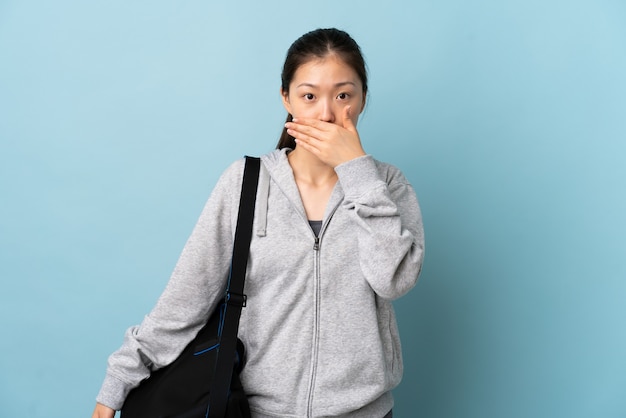 Young sport Chinese  woman with sport bag over isolated blue wall covering mouth with hand