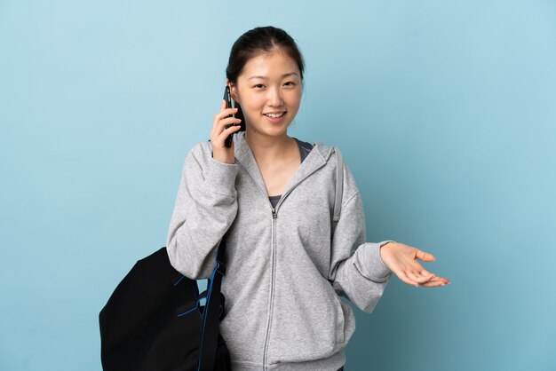 Young sport Chinese  woman with sport bag over isolated blue wall keeping a conversation with the mobile phone with someone