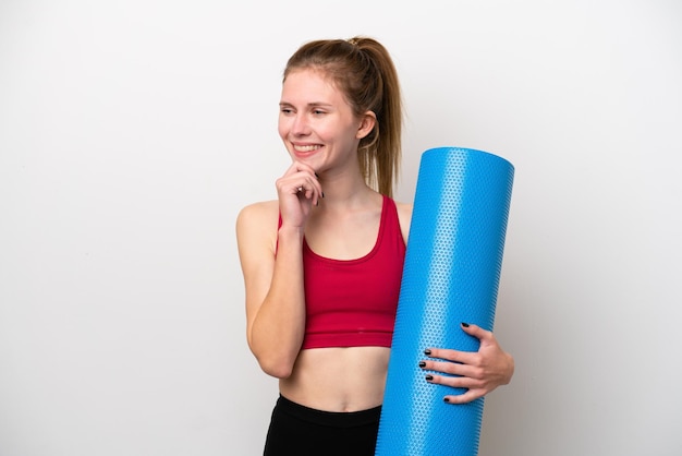 Young sport English woman going to yoga classes while holding a mat isolated on white background looking to the side and smiling