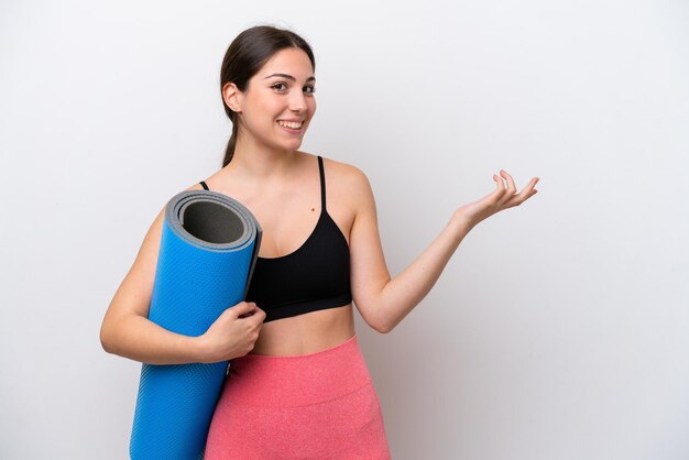 Young sport girl going to yoga classes while holding a mat isolated on white background extending hands to the side for inviting to come