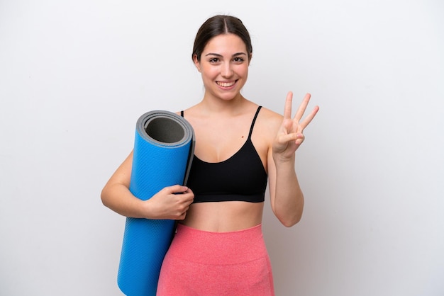 Young sport girl going to yoga classes while holding a mat isolated on white background happy and counting three with fingers