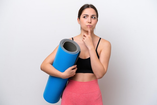 Young sport girl going to yoga classes while holding a mat isolated on white background having doubts while looking up