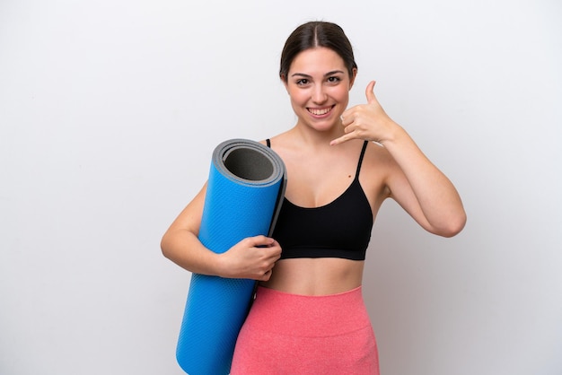 Young sport girl going to yoga classes while holding a mat isolated on white background making phone gesture Call me back sign