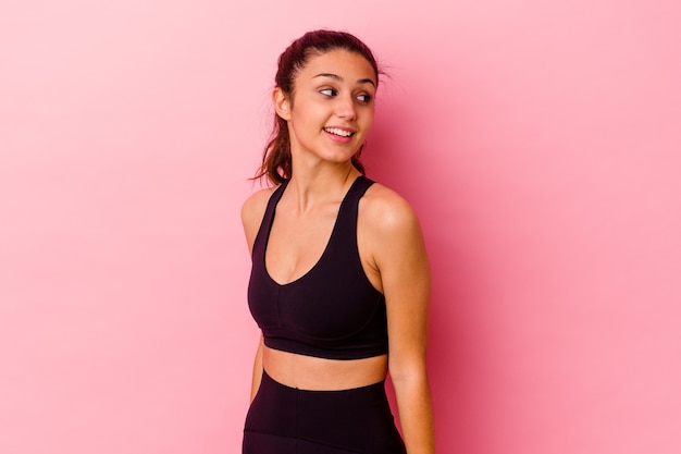 Young sport Indian woman isolated on pink wall looks aside smiling, cheerful and pleasant.