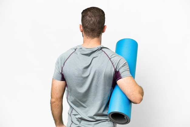 Young sport man going to yoga classes while holding a mat over isolated white background in back position