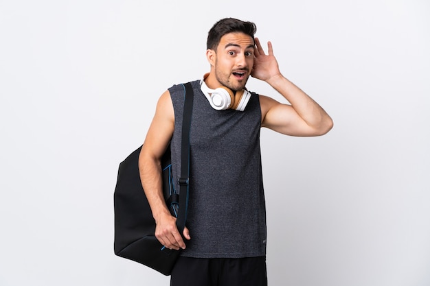 Young sport man with sport bag isolated on white background listening to something by putting hand on the ear