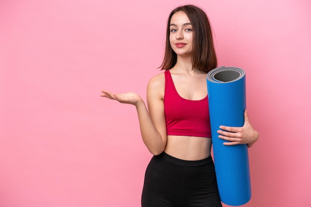 Young sport Ukrainian woman going to yoga classes while holding a mat isolated on pink background extending hands to the side for inviting to come