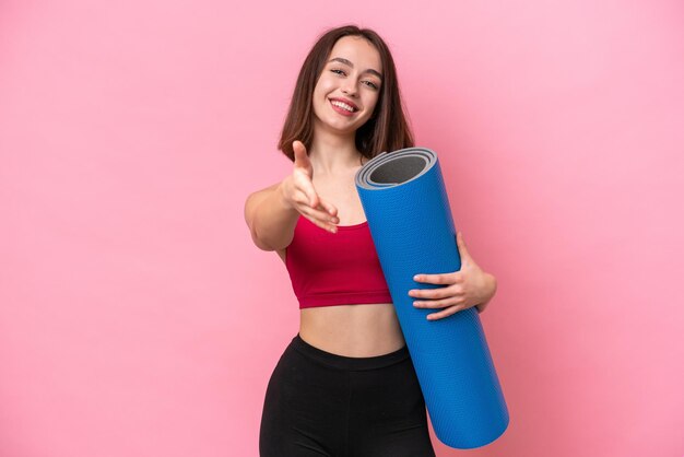 Young sport Ukrainian woman going to yoga classes while holding a mat isolated on pink background shaking hands for closing a good deal