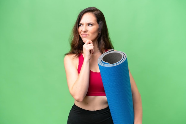 Young sport woman going to yoga classes while holding a mat over isolated background having doubts