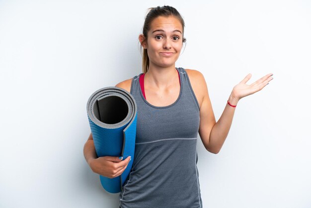 Young sport woman going to yoga classes while holding a mat isolated on white background having doubts while raising hands