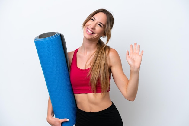 Young sport woman going to yoga classes while holding a mat isolated on white background saluting with hand with happy expression