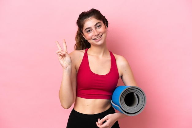 Young sport woman going to yoga classes while holding a mat smiling and showing victory sign