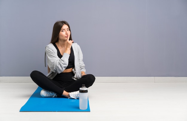 Young sport woman sitting on the floor with mat doing silence gesture