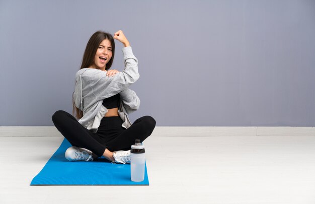 Young sport woman sitting on the floor with mat making strong gesture