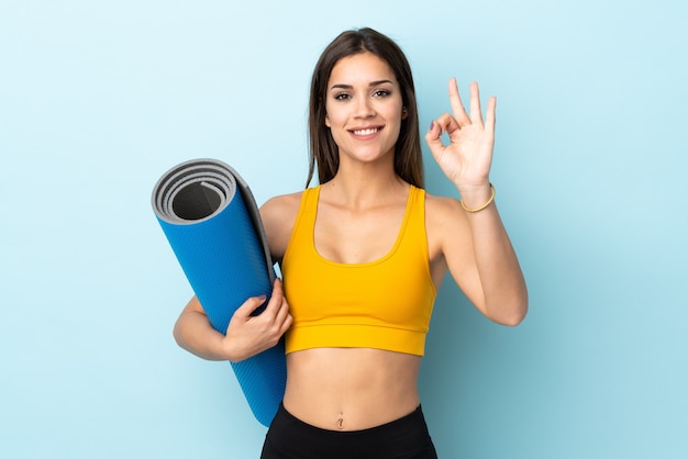 Young sport woman with mat on blue showing ok sign with fingers