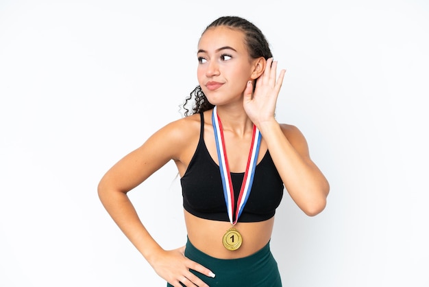 Young sport woman with medals isolated on white background listening to something by putting hand on the ear