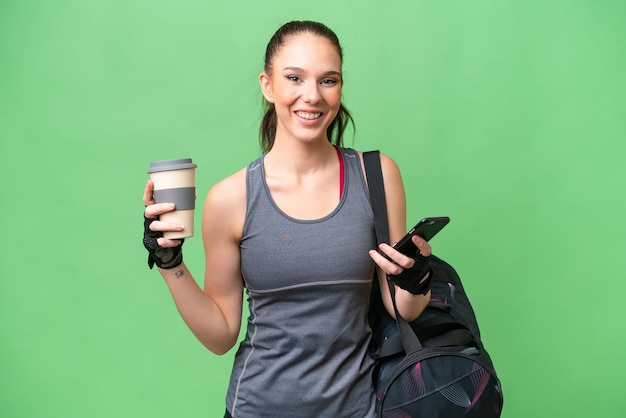 Photo young sport woman with sport bag over isolated background holding coffee to take away and a mobile