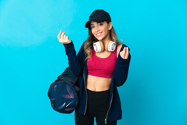 Young sport woman with sport bag isolated on blue wall making money gesture