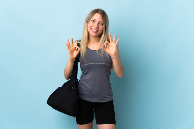 Young sport woman with sport bag isolated on blue wall showing ok sign with two hands