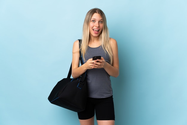 Young sport woman with sport bag isolated on blue wall surprised and sending a message