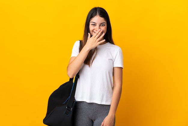 Young sport woman with sport bag isolated on yellow happy and smiling covering mouth with hand