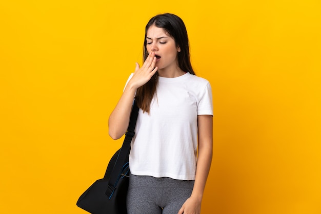 Young sport woman with sport bag isolated on yellow yawning and covering wide open mouth with hand