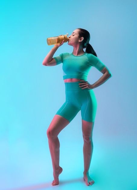 Young sportive woman training and drinking water, isolated on gradient studio background in neon light