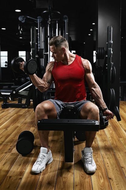 Young sports athlete fitness model in the gym shakes his biceps with dumbbells. Sports motivation, low key, high contrast. The concept of a healthy lifestyle, life movement, copy space.