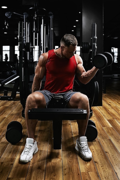 Young sports athlete fitness model in the gym shakes his biceps with dumbbells. Sports motivation, low key, high contrast. The concept of a healthy lifestyle, life movement, copy space.