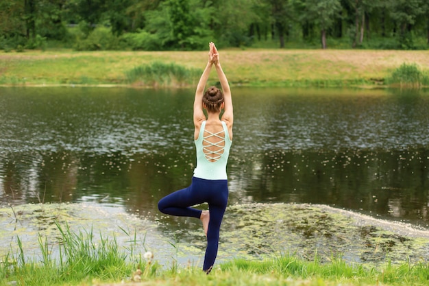 A young sports girl practices yoga on a green lawn by the river, yoga assans posture. Meditation and unity with nature