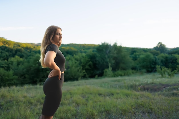 A young sportswoman in a black tracksuit keeps her hands on her belt and looks away at the sunset in the park