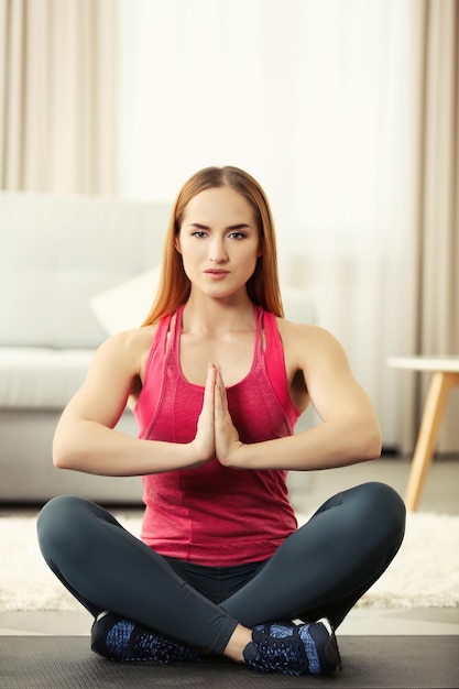 Young sportswoman sitting in lotus position on a mat at home