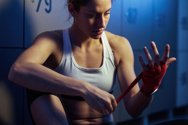 Young sportswoman wrapping hands with boxing bandage while preparing for workout in locker room