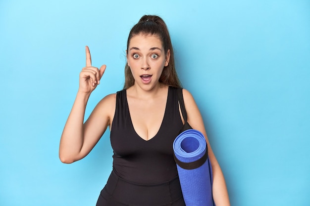 Young sporty Caucasian woman with yoga mat in blue studiohaving an idea inspiration concept