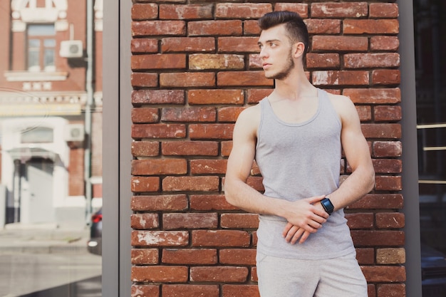 Young sporty man using smart watch, standing at brick wall background, copy space. Modern technology and fitness concept