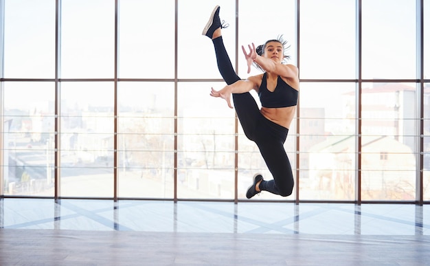 Young sporty woman in sportswear jumping and doing athletic tricks against window in the air.