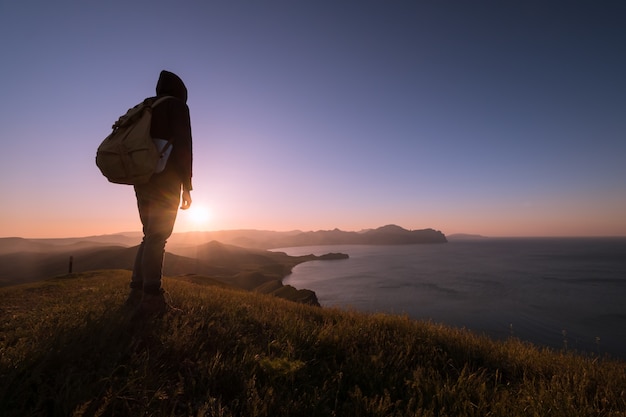 Young standing man with backpack. Hiker on the stone on the seashore at colorful sunset sky. Beautiful landscape with sporty man rocks sea and clouds at sunset. Sporty lifestyle